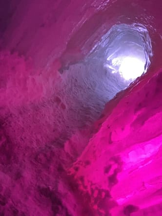 Tunnel carved into ice with snow on the ground on bright pink lights shining around the ice. 